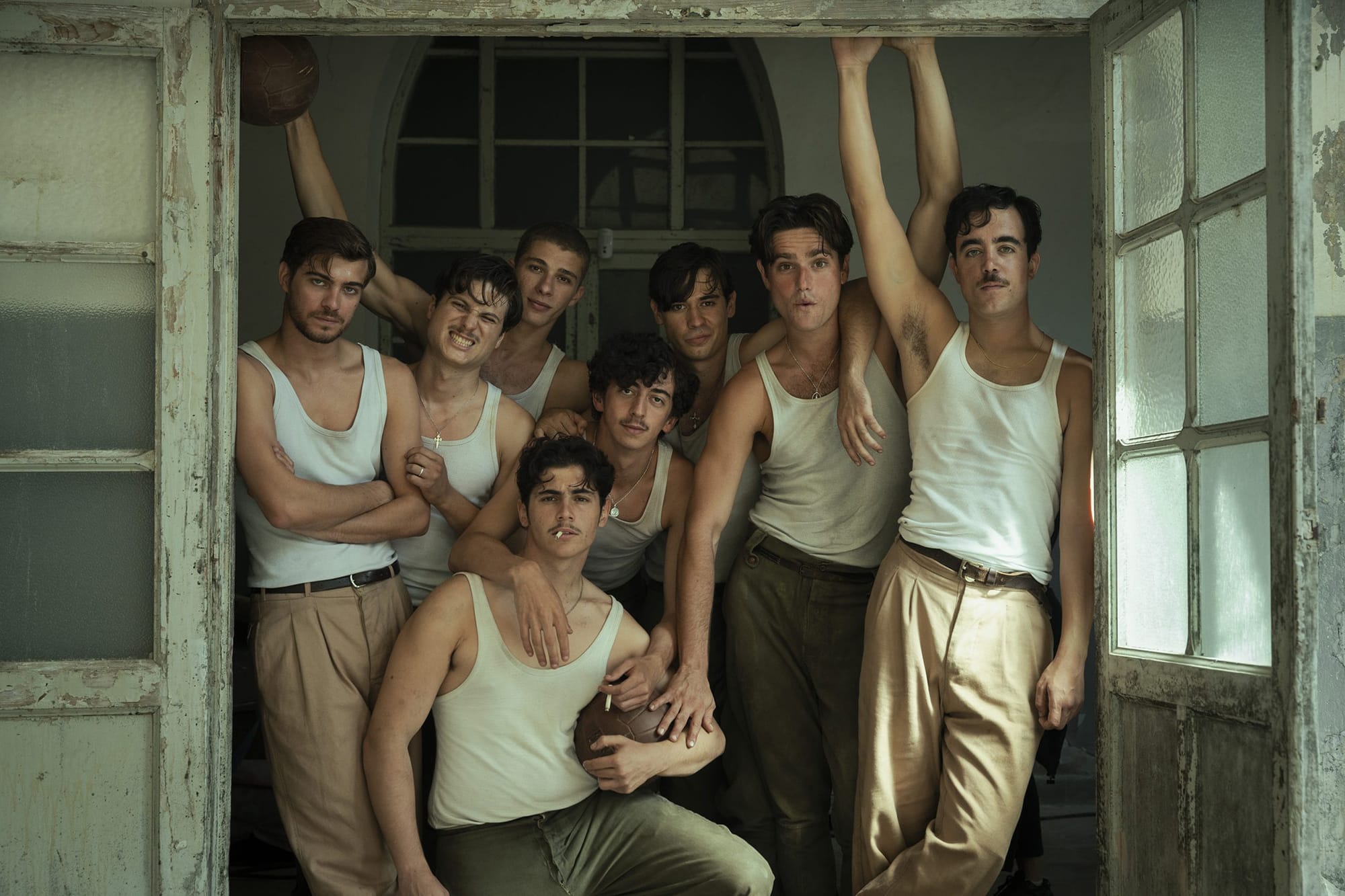 Fotografía promocional de la película 'La bola negra' que muestra a un grupo de ocho actores jóvenes vestidos con ropa de época, incluyendo camisetas blancas de tirantes, posando juntos en un espacio rústico junto a unos grandes ventanales. Uno de ellos sostiene un balón antiguo de cuero oscuro.
