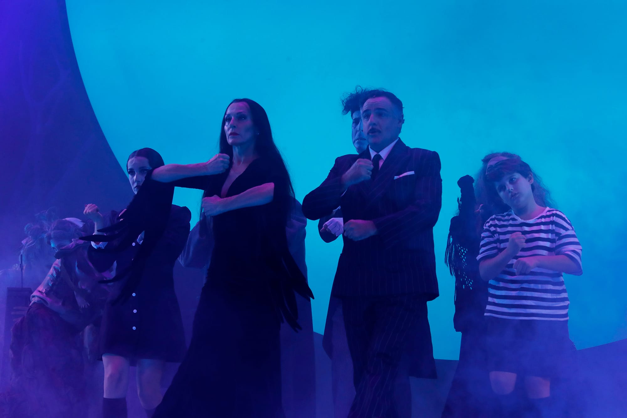 Escena del musical donde los actores que interpretan a Morticia y Gómez Addams ejecutan un paso de baile rodeados del resto de la familia bajo una espesa niebla y focos azules.