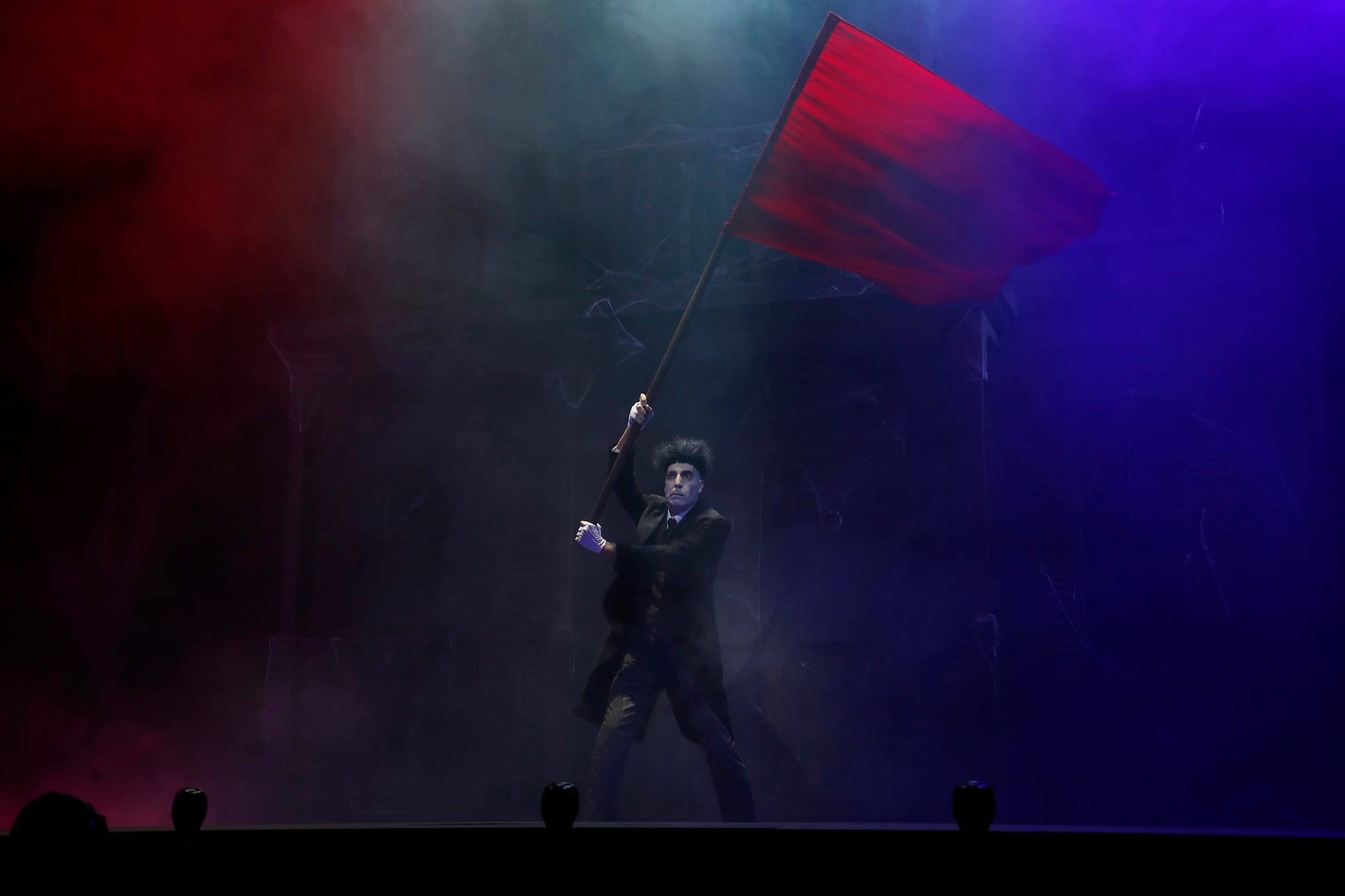 El actor Javier Canales vestido con el traje clásico del mayordomo Lurch, sosteniendo y ondeando una gran bandera roja sobre el escenario del Teatro Calderón.