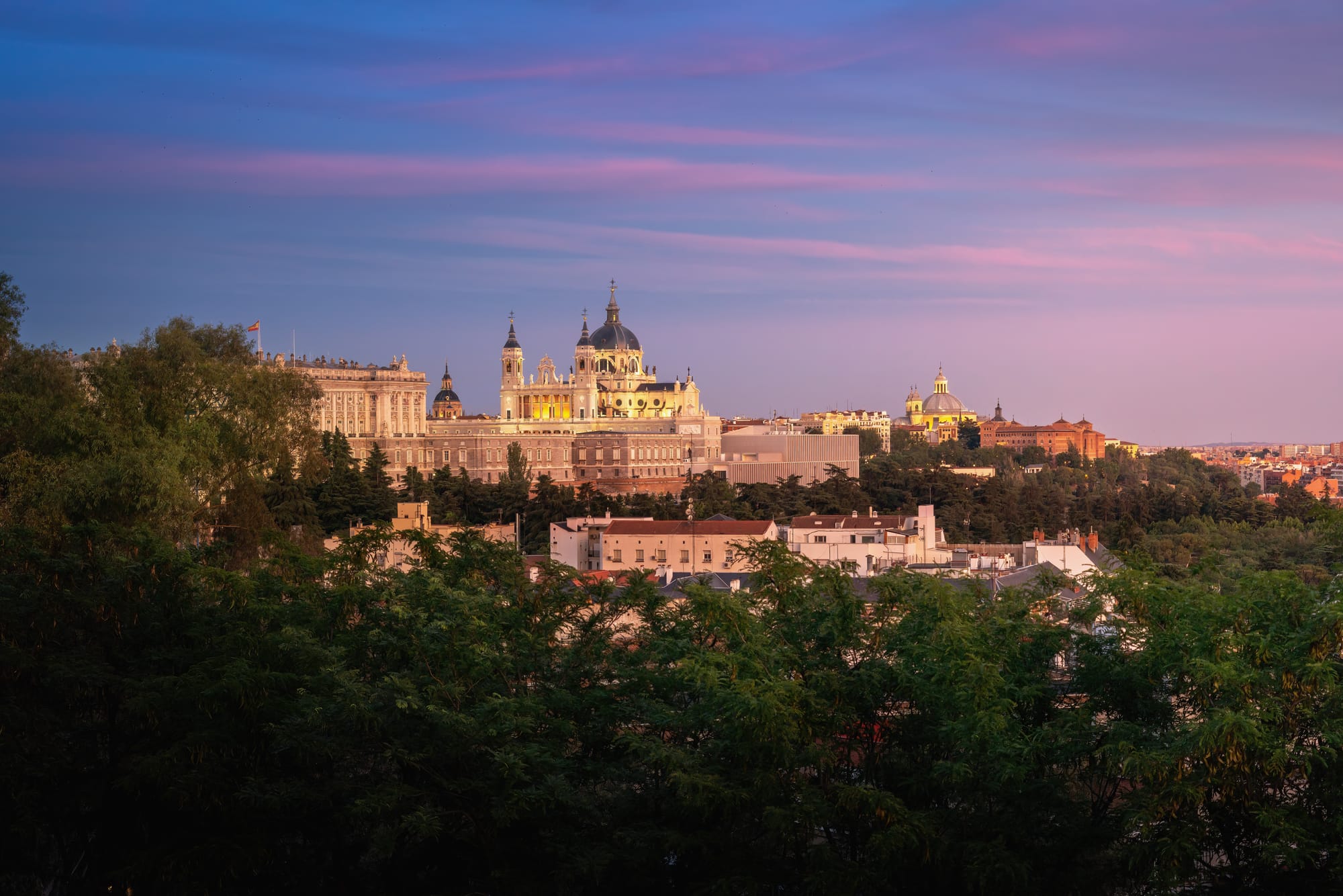 Vista panorámica de Madrid al atardecer con el cielo teñido de tonos rosados y azules, destacando la Catedral de la Almudena y el Palacio Real sobre los árboles.