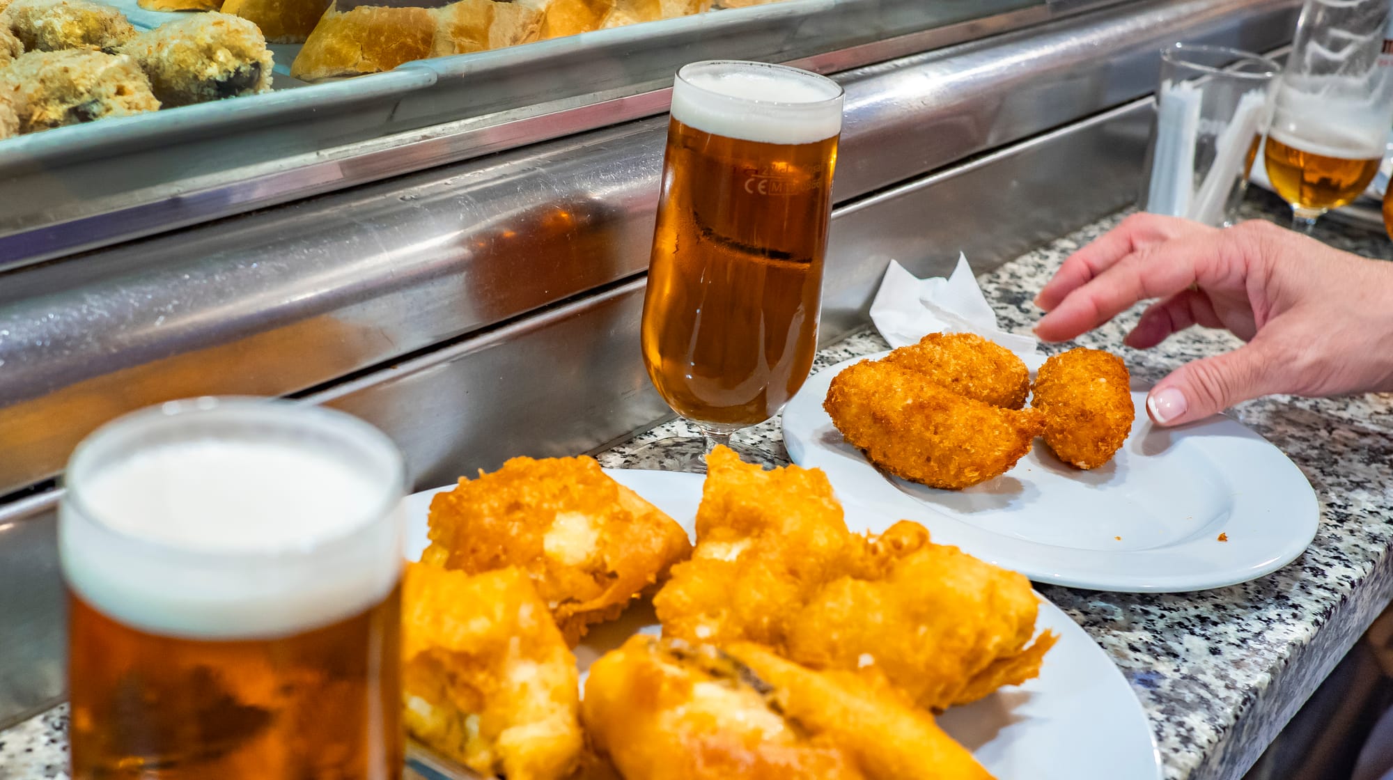 Primer plano de la barra de mármol de un bar con copas de cerveza y platos de tapas fritas, mientras la mano de un cliente coge una croqueta.