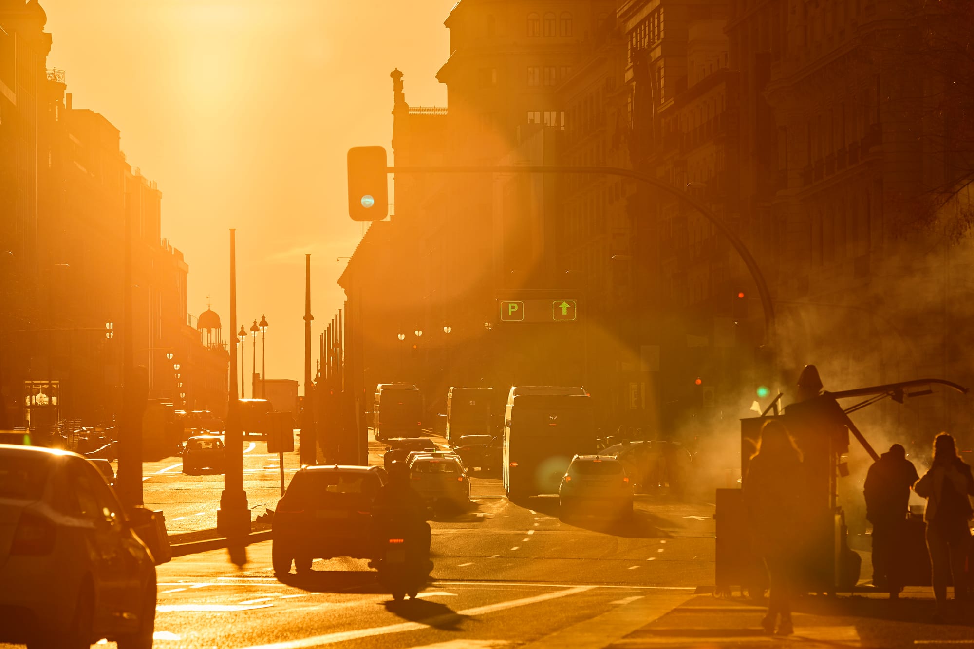 Fotografía urbana de una calle de Madrid bañada por la intensa luz dorada y anaranjada del sol del atardecer, mostrando siluetas a contraluz de coches y peatones.