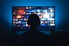 Persona sosteniendo un mando a distancia frente a un televisor que muestra el menú de una plataforma de streaming.