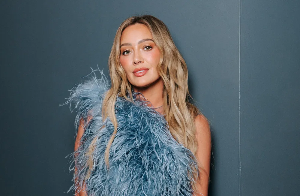 Retrato de estudio de Hilary Duff mirando a cámara, luciendo un llamativo top azul de plumas frente a un fondo liso oscuro.